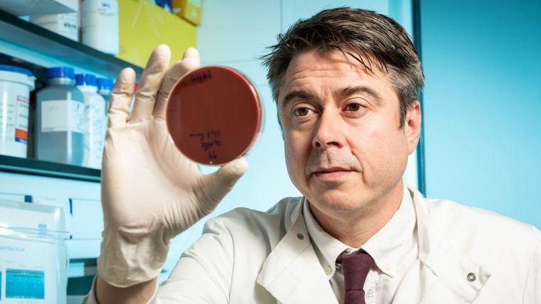Professor Sam Sheppard in lab coat, holding up a petri-dish, in a laboratory