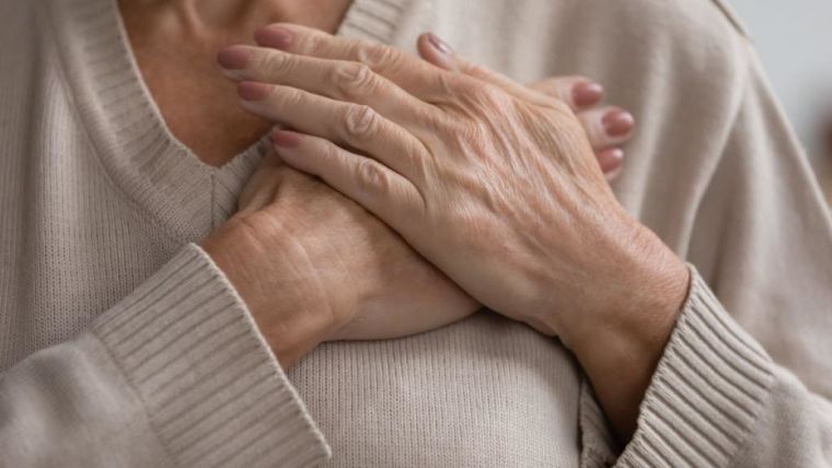 Older lady wearing a beige sweater, holding her hands crossed over her heart